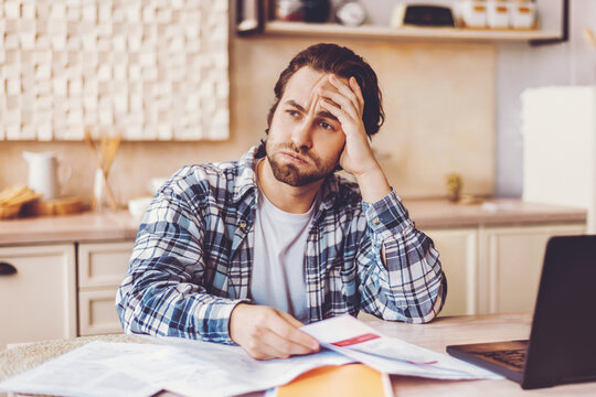 A young man sits at a kitchen table with a laptop and stacks of papers. He looks worried while reviewing bills and other financial documents. It appears to be daytime in a simple kitchen