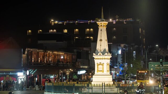 Nighttime scene with illuminated town center. Busy street with vehicles and pedestrians. Tall structure stands prominently in the middle. Vibrant lights reflect off nearby buildings and signage
