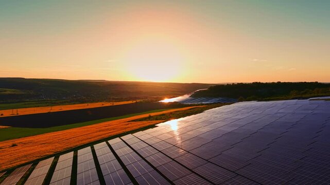 A view of solar panels set in a rural area with fields and hills during sunrise. The sun rises in the background, creating a warm glow over the landscape and the panels.