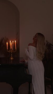 Tall blonde woman in a long off-shoulder white dress leans with both hands on a dark polished grand piano. A brass candelabrum with five lit candles sits on