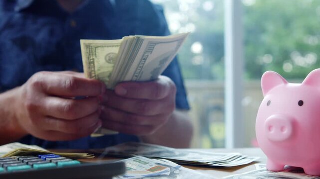 A man sits comfortably at a table in his home, meticulously counting and organizing his dollars while placing them next to a cute pink piggy bank