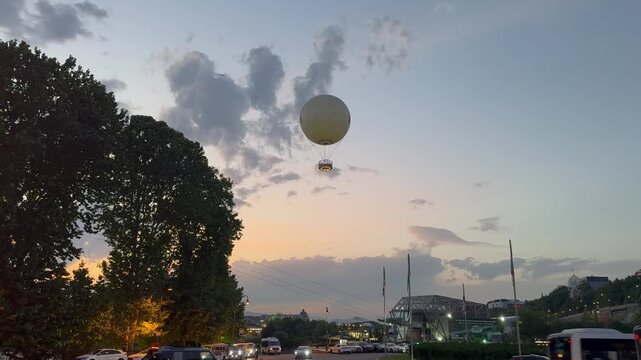 A large white tethered balloon floats gracefully in the evening sky at sunset, while city traffic moves below along a busy street with lush green trees in Tbilisi.

