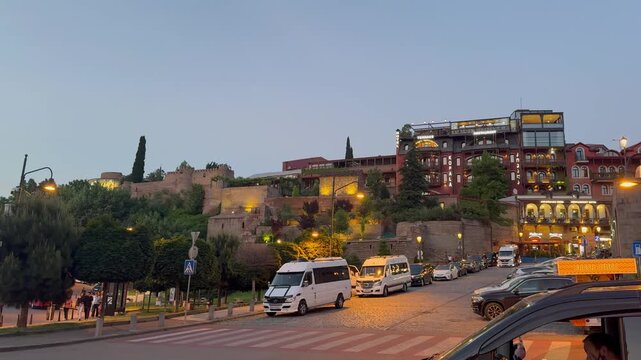 Scenic evening view of Tbilisi old town in Georgia, featuring the illuminated Narikala fortress on the hill, Metekhi church, passing cable cars, and busy street traffic at sunset.

