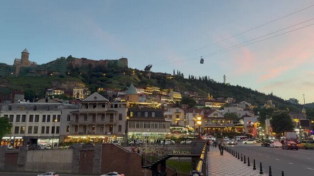 Scenic evening view of Tbilisi old town in Georgia, featuring the illuminated Narikala fortress on the hill, Metekhi church, passing cable cars, and busy street traffic at sunset.

