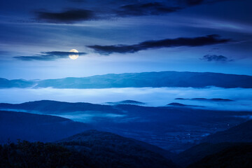 Fototapeta premium foggy carpathian countryside scenery in summer at night. mysterious view of mountainous landscape with rural fields on rolling hills. full moon over fog in the valley. clouds above the mountain