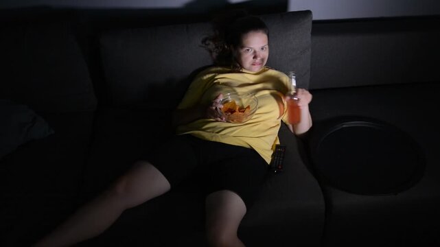 Young overweight woman lying on a sofa at night with a sad expression, slowly eating tortilla chips and switching channels with a remote control. 