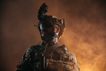 Fototapeta premium Low angle close up portrait of a tactical soldier in camouflage uniform and night vision helmet standing against dramatic orange smoke, representing special operations