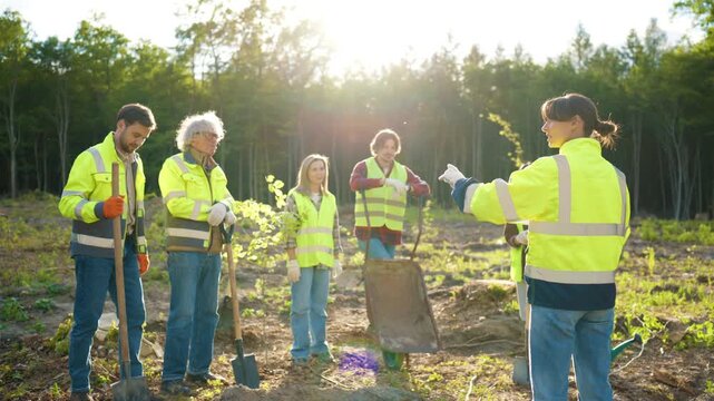Environmental team receiving instructions before tree planting project. Diverse volunteers wearing safety jackets and holding tools. Eco restoration work on land. Sustainable forestry initiative.
