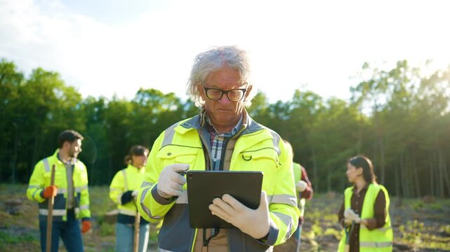 Adult Caucasian environmental specialist reviewing restoration progress on tablet at reforestation site. Elderly volunteer monitoring ecology project after logging. Sustainable forest recovery.