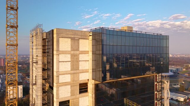 Aerial orbit around a glass clad high rise under construction, passing a yellow tower crane as warm sunlight reflects across the facade above the city.