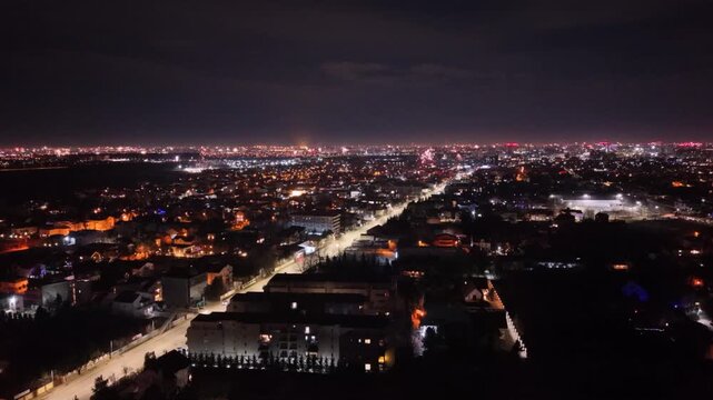 Aerial drone footage capturing New Year's Eve fireworks erupting across a brightly illuminated city at night. Multiple fireworks displays light up the urban skyline while a long boulevard stretches th
