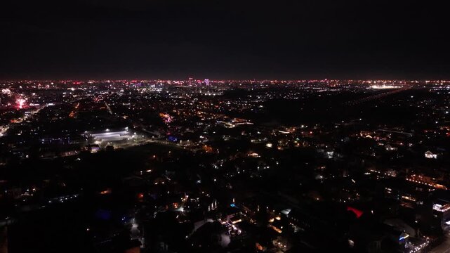 Aerial drone footage capturing New Year's Eve fireworks erupting across a brightly illuminated city at night. Multiple fireworks displays light up the urban skyline while a long boulevard stretches th