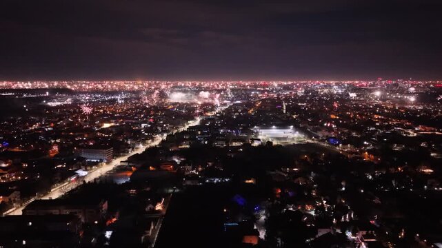 Aerial drone footage capturing New Year's Eve fireworks erupting across a brightly illuminated city at night. Multiple fireworks displays light up the urban skyline while a long boulevard stretches th