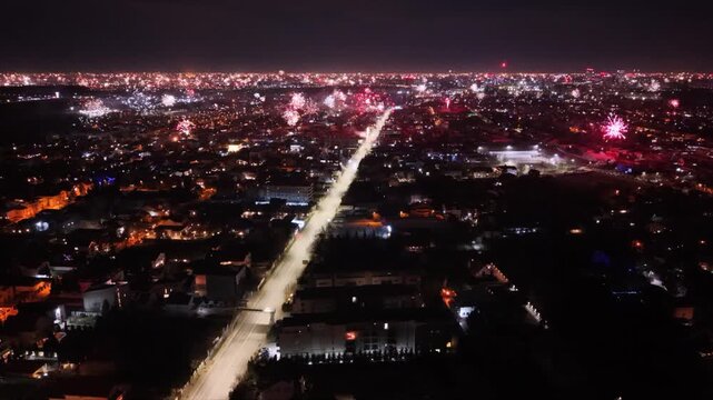 Aerial drone footage capturing New Year's Eve fireworks erupting across a brightly illuminated city at night. Multiple fireworks displays light up the urban skyline while a long boulevard stretches th