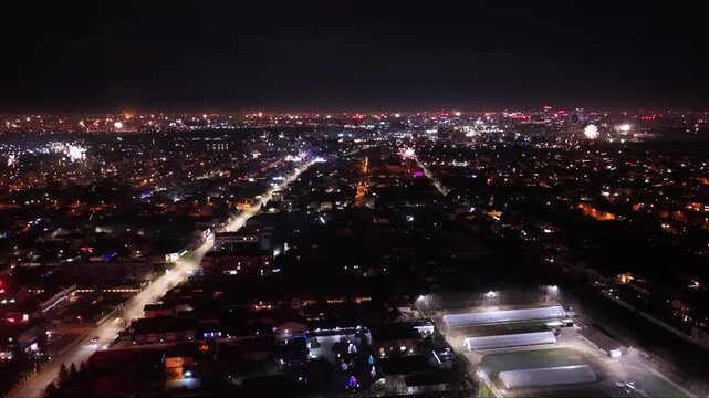 Aerial drone footage capturing New Year's Eve fireworks erupting across a brightly illuminated city at night. Multiple fireworks displays light up the urban skyline while a long boulevard stretches th