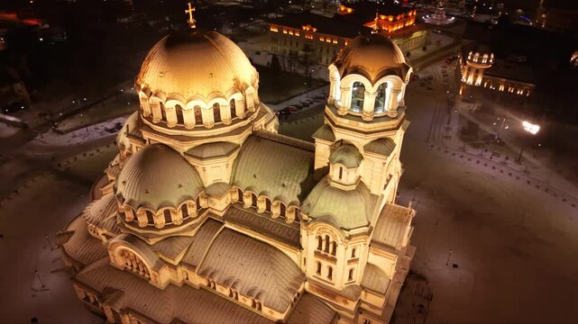 Night aerial view of a brightly lit cathedral with golden domes, surrounded by snowy grounds and city streets of Sofia, Bulgaria. Orthodox Christian church seen from above at night