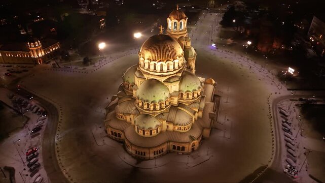 Night aerial view of a brightly lit cathedral with golden domes, surrounded by snowy grounds and city streets of Sofia, Bulgaria. Orthodox Christian church seen from above at night