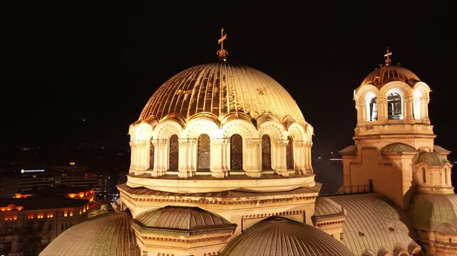 Night aerial view of a brightly lit cathedral with golden domes, surrounded by snowy grounds and city streets of Sofia, Bulgaria. Orthodox Christian church seen from above at night