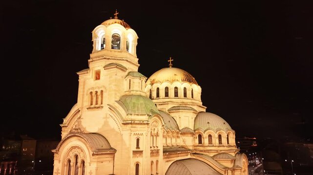 Cinematic aerial drone footage of the illuminated Alexander Nevsky Cathedral on a snowy winter night in Sofia, Bulgaria. The golden domes glow against the dark cityscape while fresh snow blankets the 