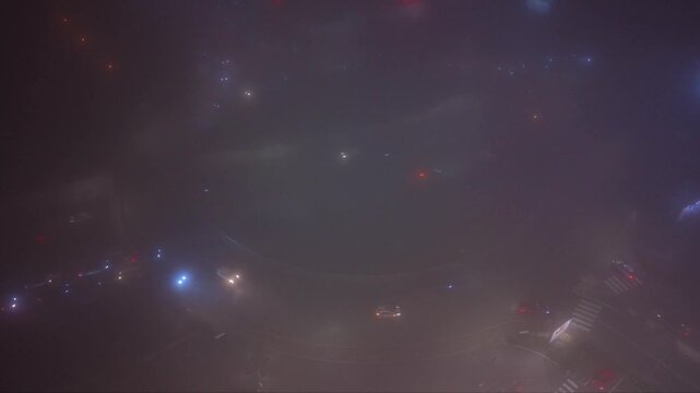 Atmospheric aerial drone footage of a city at night covered by dense fog with vehicle headlights and illuminated signs glowing through the thick mist. The low visibility creates a mysterious urban lan