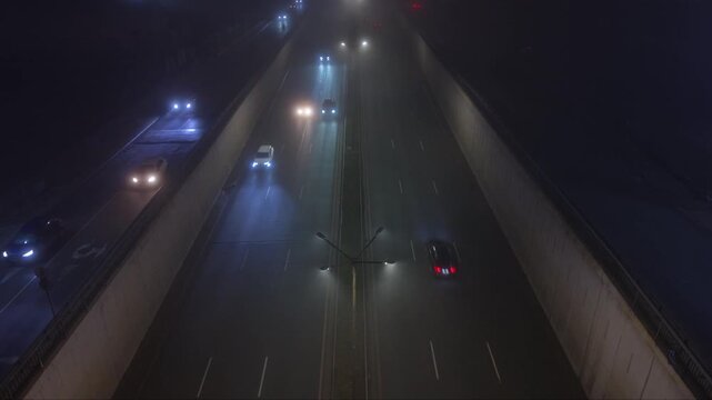 Atmospheric aerial drone footage of a city at night covered by dense fog with vehicle headlights and illuminated signs glowing through the thick mist. The low visibility creates a mysterious urban lan