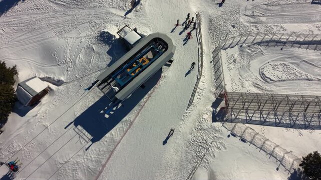 Vertical aerial drone footage of skiers and snowboarders gathering at the intersection of ski runs in a busy mountain resort. Long winter shadows stretch across freshly groomed snow as visitors prepar