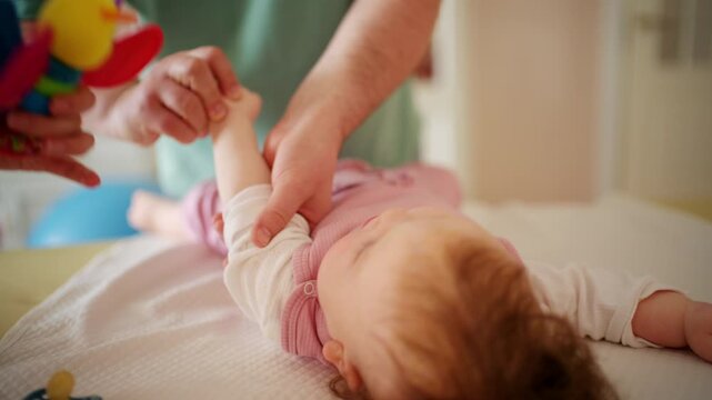 A colorful baby rattle is gently presented to an infant, while caregiver is performing mobility exercises