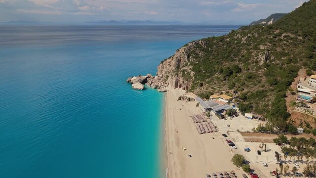 A breathtaking aerial pullback revealing a long sandy beach, dramatic coastal cliffs, and crystal-clear turquoise waters. As the drone flies backward, the scale of the coastline unfolds, showcasing th