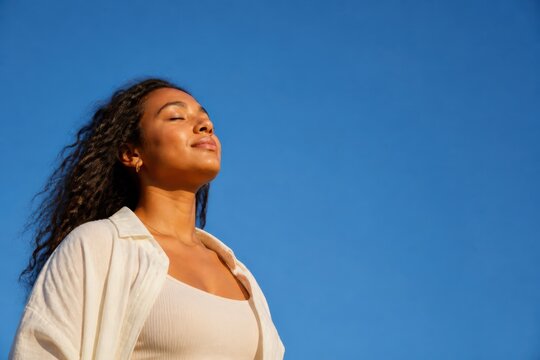 Serene young woman with eyes closed enjoying sunlight against a clear blue sky outdoors