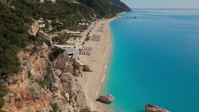 Spectacular drone flight along dramatic coastal cliffs overlooking a stunning sandy beach and vibrant turquoise waters. The aerial perspective reveals the scale of the coastline, crystal-clear sea, ro