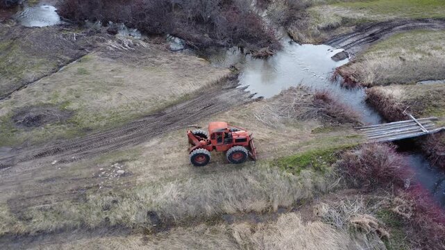 Drone orbit footage of an old red Timberjack skidder parked beside a rustic log bridge, muddy trail, and flowing spring stream in a quiet rural landscape.