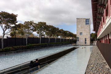 Fototapeta premium A couple walks by a tranquil water feature in front of a modern building with traditional architectural elements at Fo Guang Shan Buddhist Temple, Auckland, New Zealand.