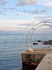 Obraz premium Minimalist metal wedding arch structure standing on a concrete seaside pier against a calm blue ocean horizon. High-quality seascape photograph perfect for romantic destination wedding planning websit