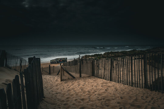 Dark low key coastal scenery in Labenne Ocean featuring a moody beach access path through sand dunes and wooden fences under an overcast sky, solitude landscape in Landes, France.