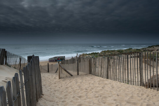 Moody coastal landscape in Labenne Ocean with a beach access path through sand dunes under a powerful dark stormy sky with heavy clouds, traditional fences in Landes, Aquitaine.