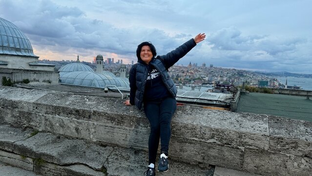 Happy woman posing on the rooftop balcony of Suleymaniye Mosque complex in Istanbul, Turkey. Ancient Ottoman empire stone buildings architecture, cloudy sky and scenic city panorama background.