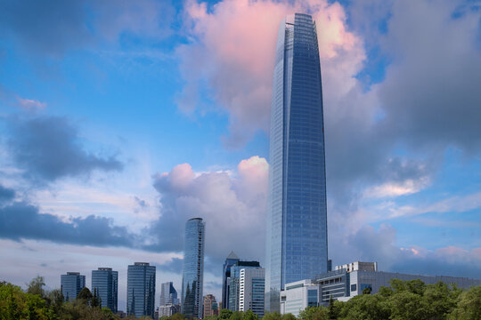 A scenic view of a modern glass skyscraper rising above the financial center skyline and green trees under a dramatic blue sky with pink tinted clouds in the capital city of Santiago Chile