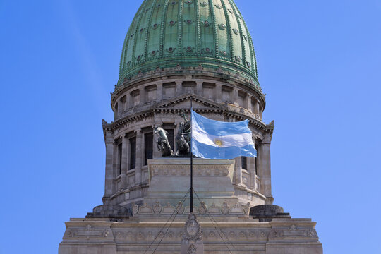 A close up view of the green copper dome and stone facade of the Palace of the Argentine National Congress featuring the national flag waving on a flagpole under a clear blue sky in Buenos Aires