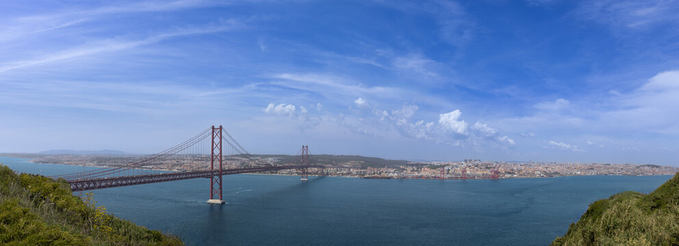 Panoramic view of the 25 de Abril suspension bridge spanning the Tagus river under a blue sky in Lisbon Portugal panorama