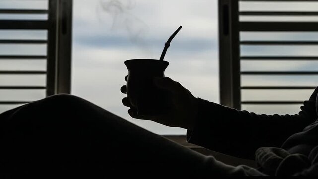 Silhouette of a person holding a steaming mate gourd by a window
