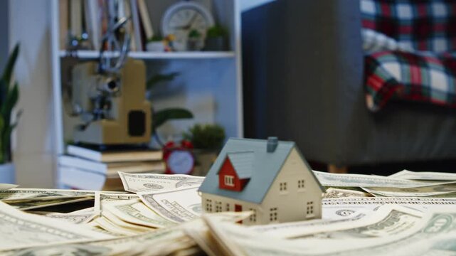 A miniature house balances on a pile of money in a warm living room. The inviting space reflects hopes for homeownership and dreams of the future