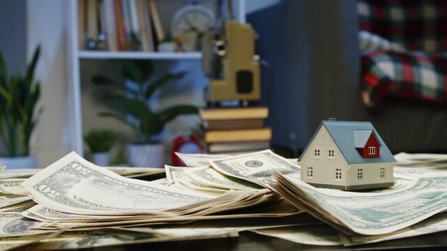A charming miniature house sits atop a pile of dollar bills, symbolizing the aspirations tied to real estate and wealth, all set in a warm, inviting living space