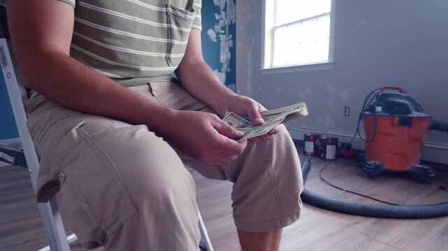 A man sits comfortably, counting dollar bills to determine how much he needs for home repairs. He thoughtfully calculates costs among tools and supplies