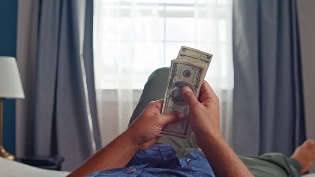 Hands eagerly count crisp dollar bills while relaxing on a bed, the warm afternoon sun streaming through the window, creating a tranquil atmosphere