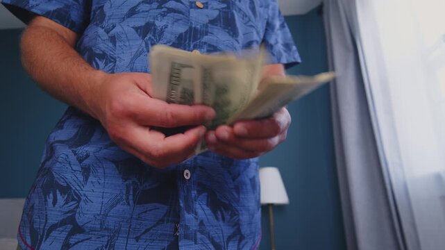 A person wearing a colorful shirt counts a stack of cash inside a bright, cozy apartment. Sunlight streams through the window, creating a warm atmosphere