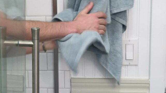 Close up of man drying hands on a blue towel hanging next to a glass shower door in bathroom