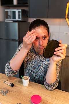 Costumed performer with beige scar wax on her brow holding a mirror at home