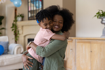 Obraz premium Mid adult African American mother and son hugging in living room with blue balloons, watch visible