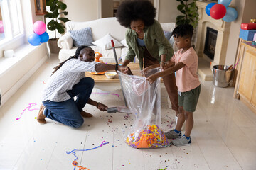 Obraz premium African American parents and child cleaning confetti in living room with clear trash bag and broom