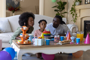 Obraz premium African American parents with son kneeling by wooden table in living room blowing out rainbow cake